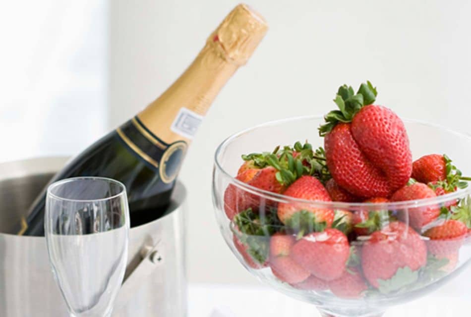 A glass bowl of strawberries next to a silver bucket with a bottle of champagne and an empty glass.