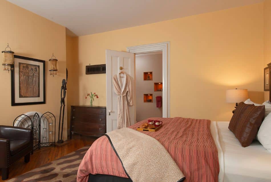 Interior of a cozy bedroom with light yellow walls, bed with pink quilt, leather club chair and robe hanging on the door