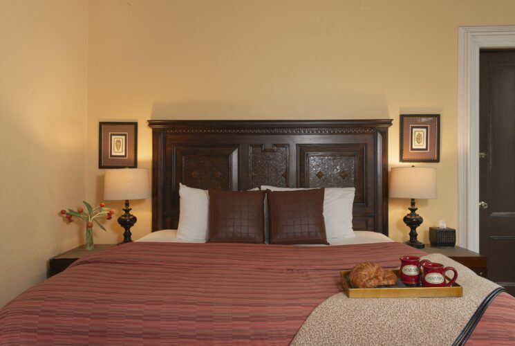 Bedroom with cream walls, ornate headboard, leather decorative pillows, tray with coffee mugs and a croissant