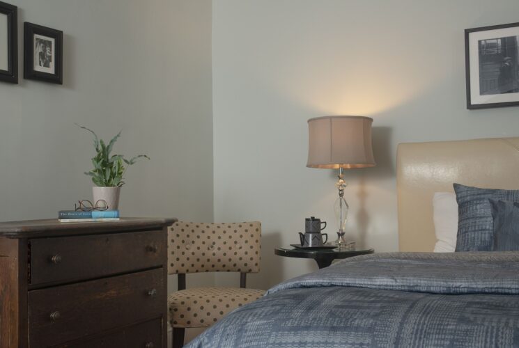 Bedroom with bed with cream headboard, side table with lamp and tea pot, sitting chair and wood dresser