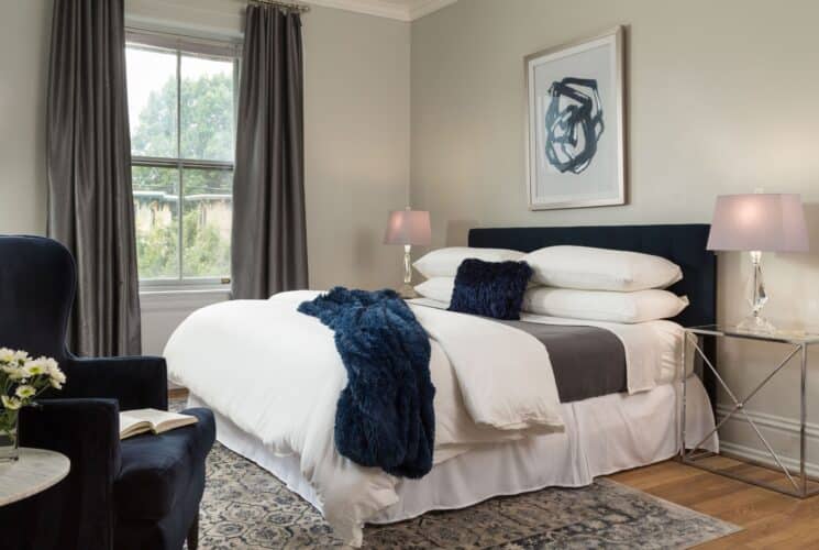 Elegant bedroom with king bed, white linens with navy pillow and throw, glass side tables with lamps, wingback chair, large window with curtains