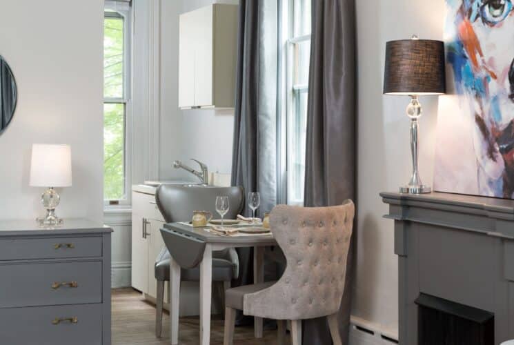 Kitchenette area of a bedroom with sink, cupboards, table with two chairs by a large window with grey curtains