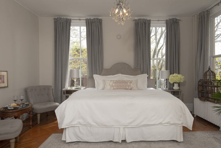 Elegant bedroom in hues of grey and white with king bed in white linens, two plush sitting chairs with coffee table, large windows with grey curtains