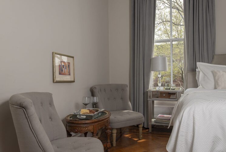 Sitting area of an elegant bedroom with two plush grey chairs, table with tray of cheese and crackers and two glasses of wine