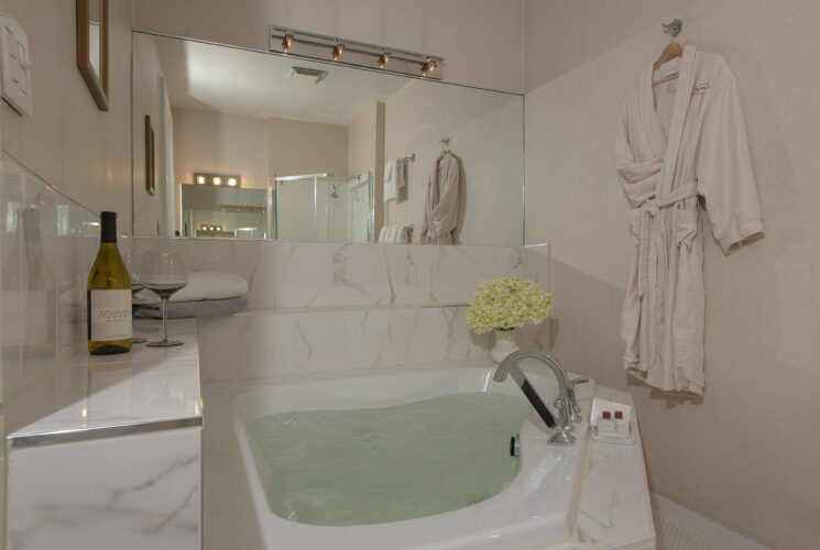 Bathroom with corner jacuzzi tub filled with water, robe hanging on a hook and bottle of wine with a glass on a marble ledge