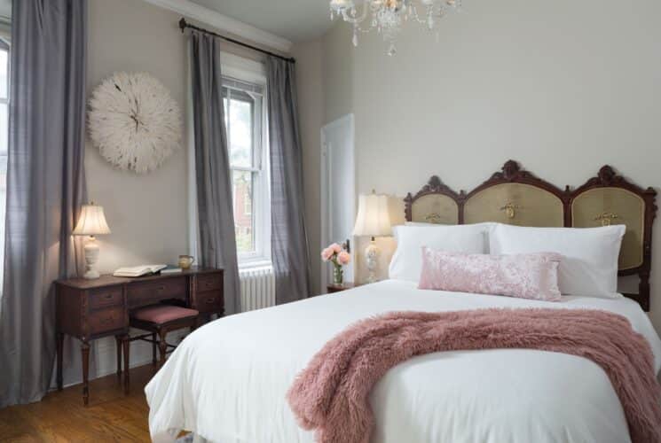 Elegant bedroom with queen bed, ornate headboard with white linens, pink pillows, side table with lamp and vase of flowers and large window with curtains