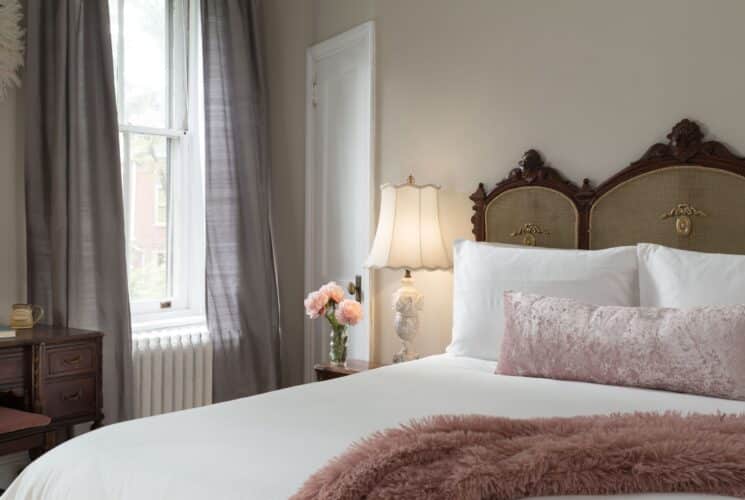 Elegant bedroom with queen bed, ornate headboard with white linens, pink pillows, side table with lamp and vase of flowers and large window with curtains