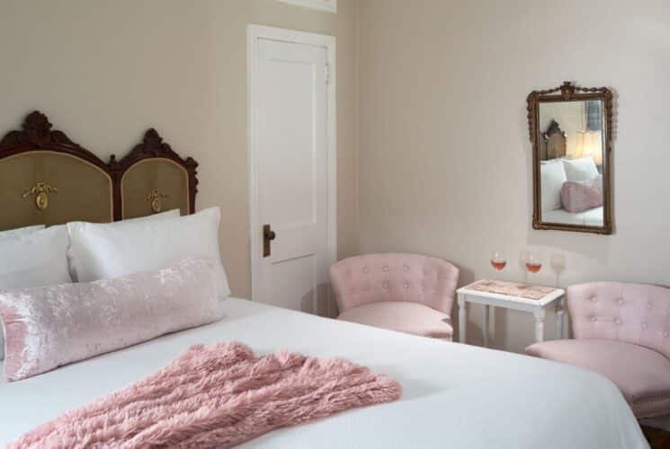 Elegant bedroom with queen bed, ornate headboard, white and pink linens, two pink sitting chairs by table with two glasses of wine