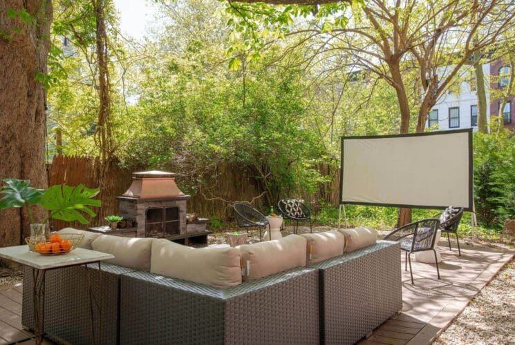 Serene fenced in backyard with tall trees, patio sectional and fireplace with seating in front of an outdoor movie screen