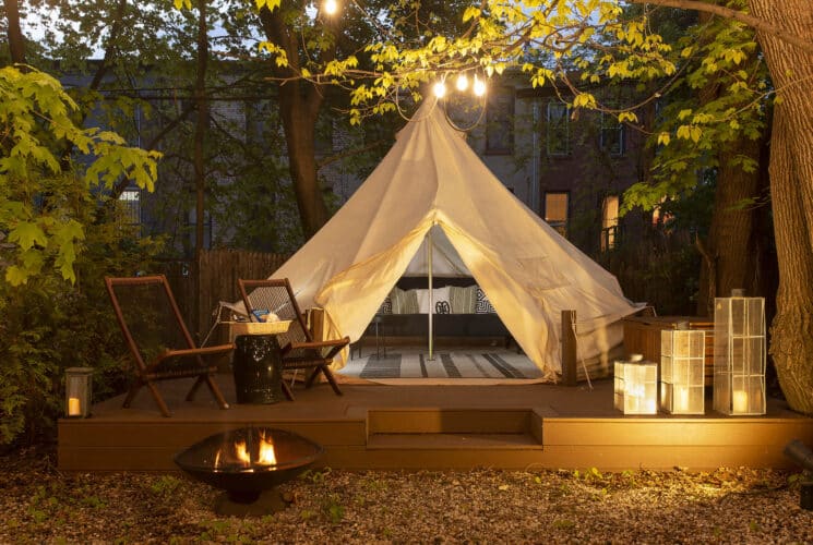 A glamping tent set up in a backyard, lit up at night with candles, a firepit, and seating