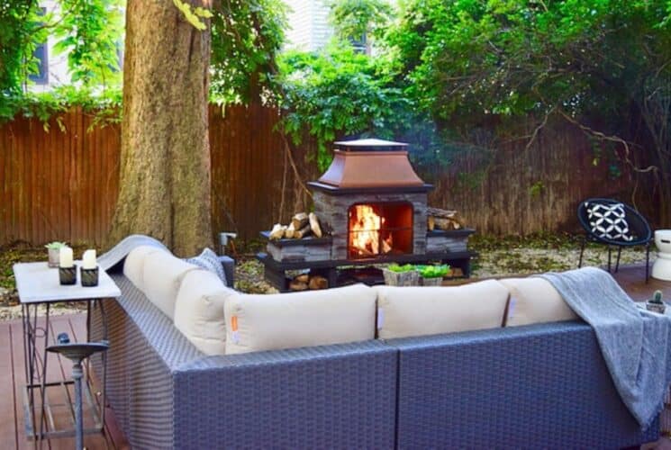 A serene fenced in backyard with patio couch with tan cushions in front of a decorative fireplace