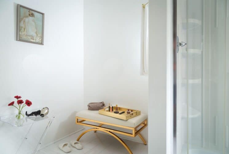 Corner of a bathroom with gold and tan bench, tray of toiletries and table with hair dryer and vase of red flowers