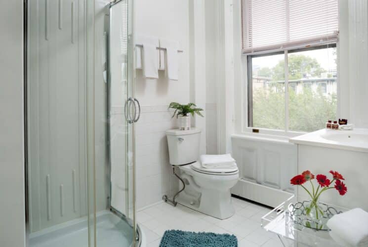 Bright white bathroom with corner glass door shower, blue rug, folded towels on rack above toilet and large window with blinds