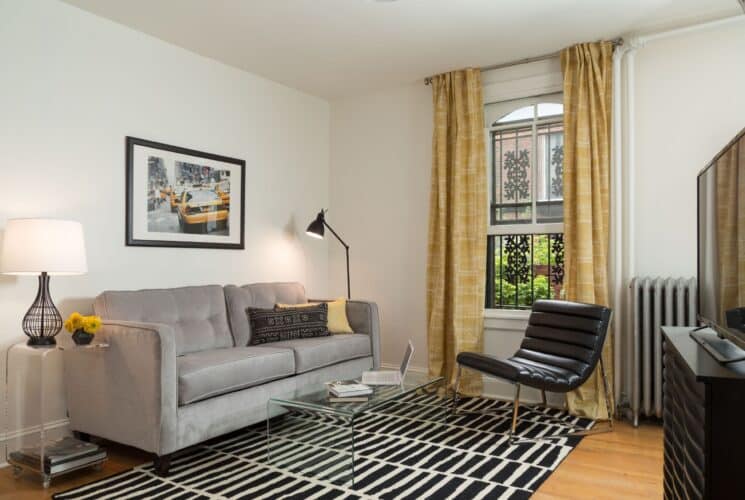 Living room with couch, black and white rug, leather chair, window with yellow curtains, dresser with TV