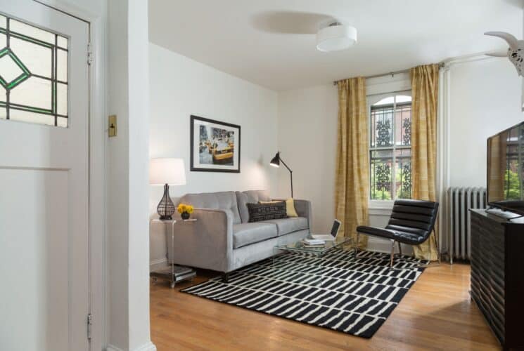 Living room with couch, black and white rug, leather chair, window with yellow curtains, dresser with TV