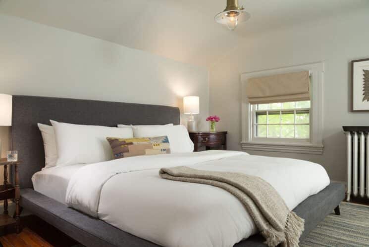 Elegant bedroom with king bed, grey headboard, white pillows and linens, window with Roman shade and side tables with lamps