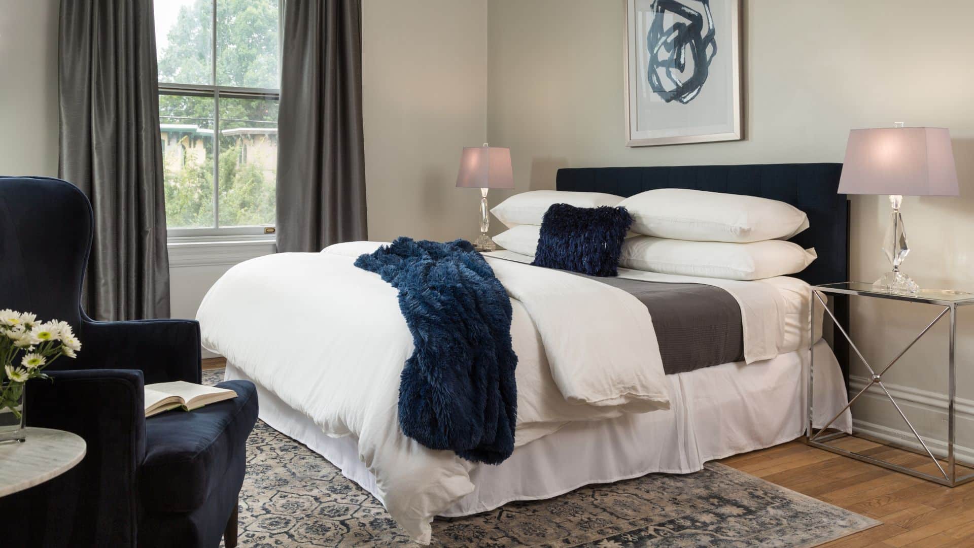 Elegant bedroom with king bed in white linens with blue blanket and pillow, wingback chair, side tables with lamps and large window with curtains