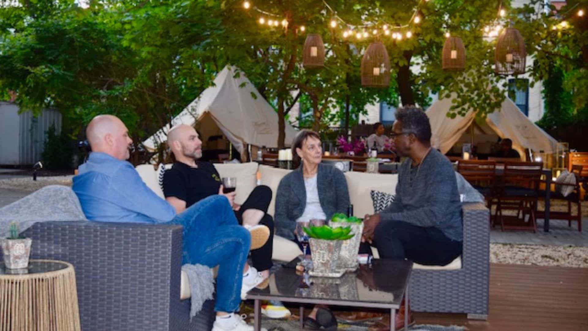 A group of people sitting on an outdoor patio sectional in a backyard with lanterns, patio lights and two glamping tents in the background.