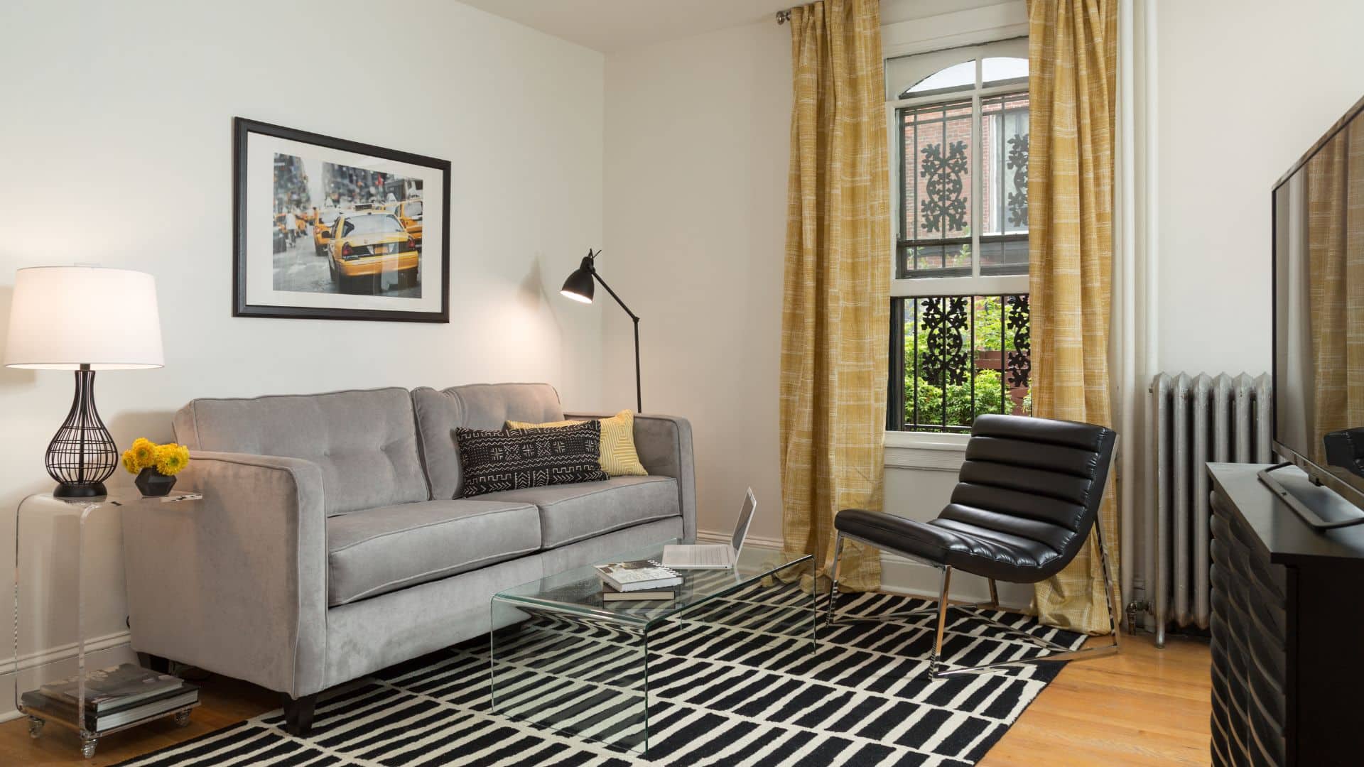 Living room with grey couch, side table with lamp, black and white rug, leather chair, dresser with TV and window with yellow curtains