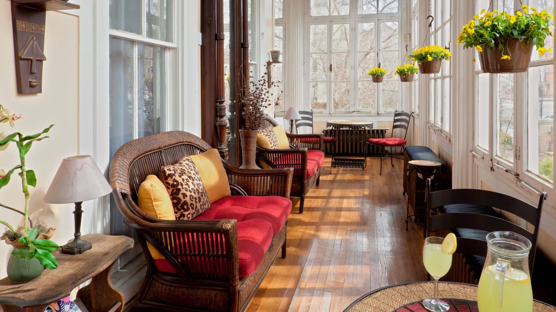 Gorgeous sunroom with walls of windows, hardwood floors, hanging flower basket, wicker furniture with red, yellow and animal print cushions