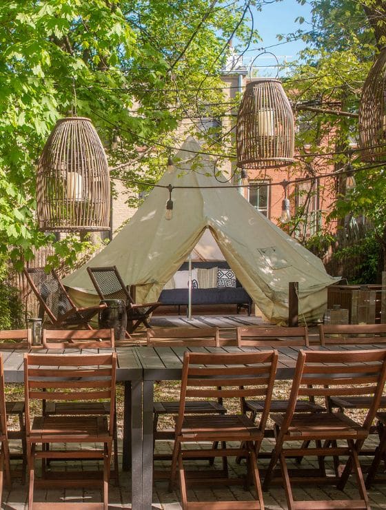 An elegant glamping test in a backyard with hanging lanterns, patio lights and a dining table with wood chairs