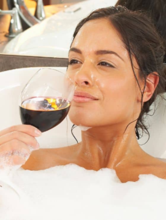 A woman with brown hair sitting a bathtub full of bubbles and holding a glass of red wine to her lips