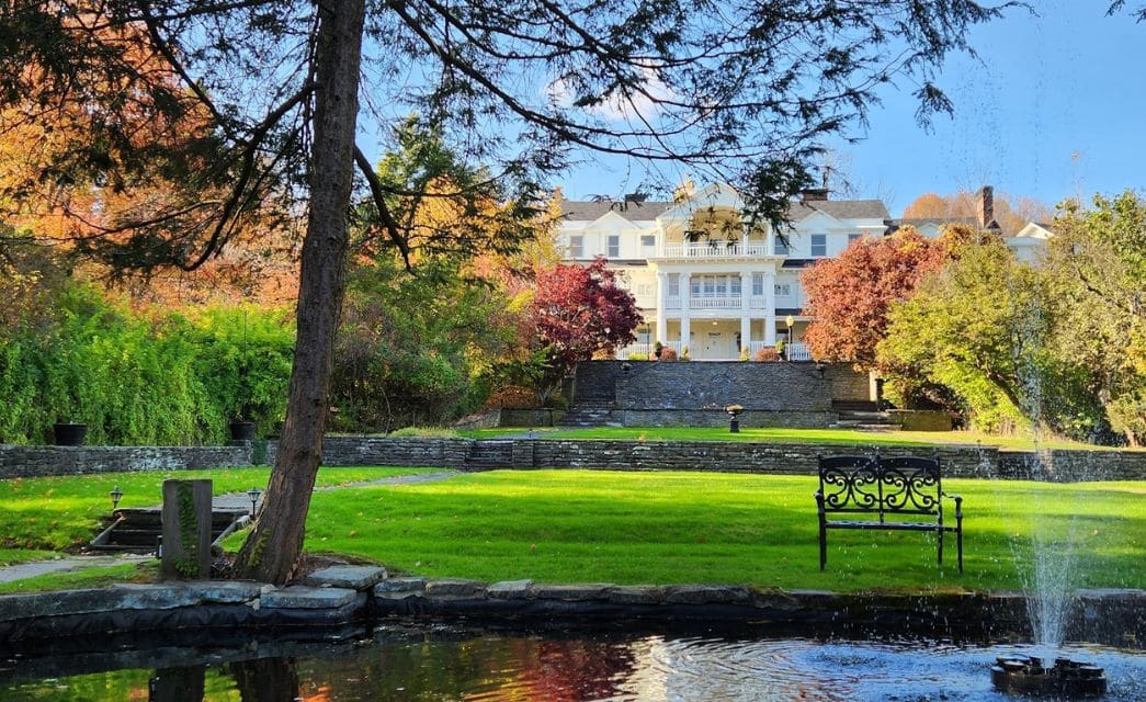 Small pond with fountain by expansive tiered lawns with a stately mansion in the background behind a stone wall