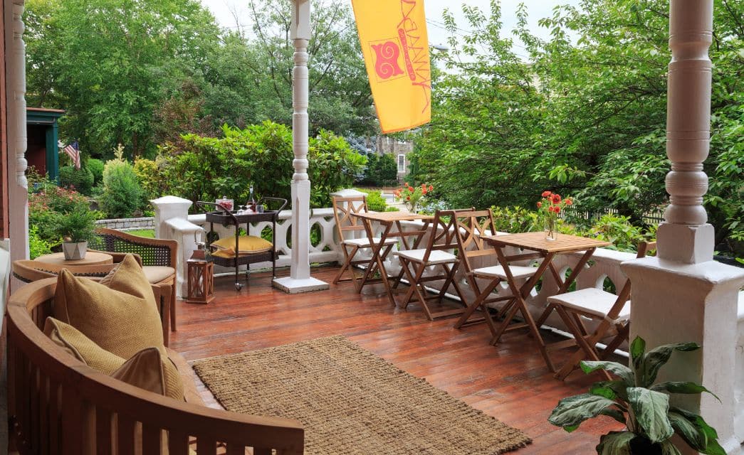 Front porch of a home with hardwood floors, two tables with chairs, decorative seating and yellow and red hanging flag