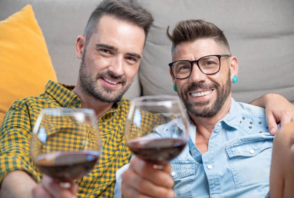 Two men sitting in front of a tan couch each holding a glass of red wine