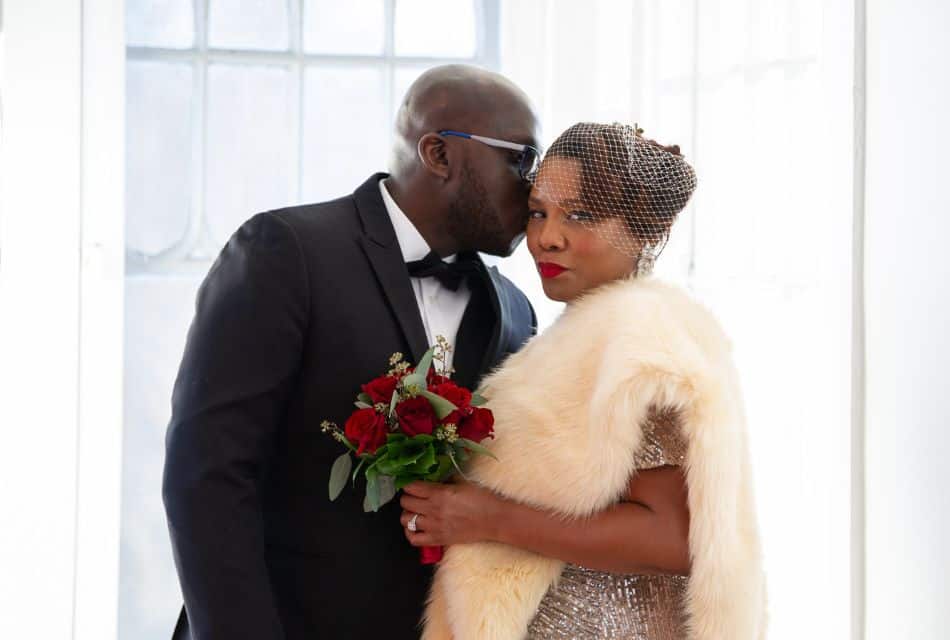 A woman in a fur shawl, gold dress and holding a bouquet of red roses next to a man in a black suit