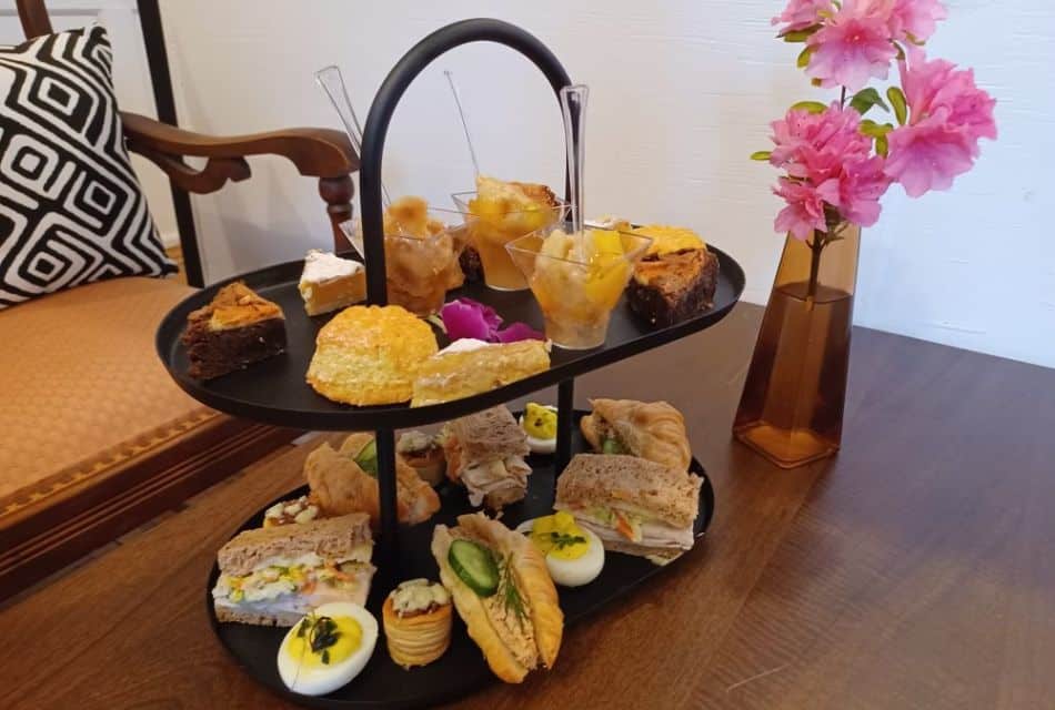 Bench seat by a window with a round marble table holding a three-tiered tray of desserts and fruit and two tea pots