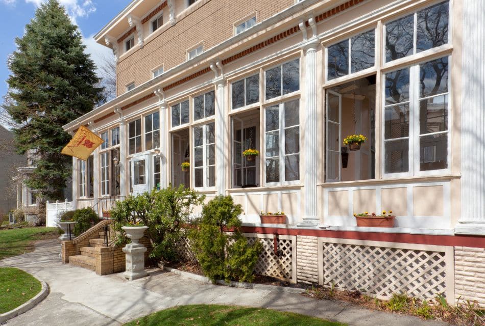 Front facade of a large home with a wall of windows, some open with hanging flower pots, bushes by front steps