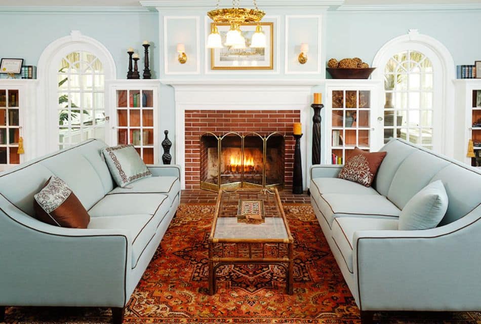 An elegant living room with two mint colored couches, glass coffee table, Oriental rug, in front of a brick fireplace with white mantle